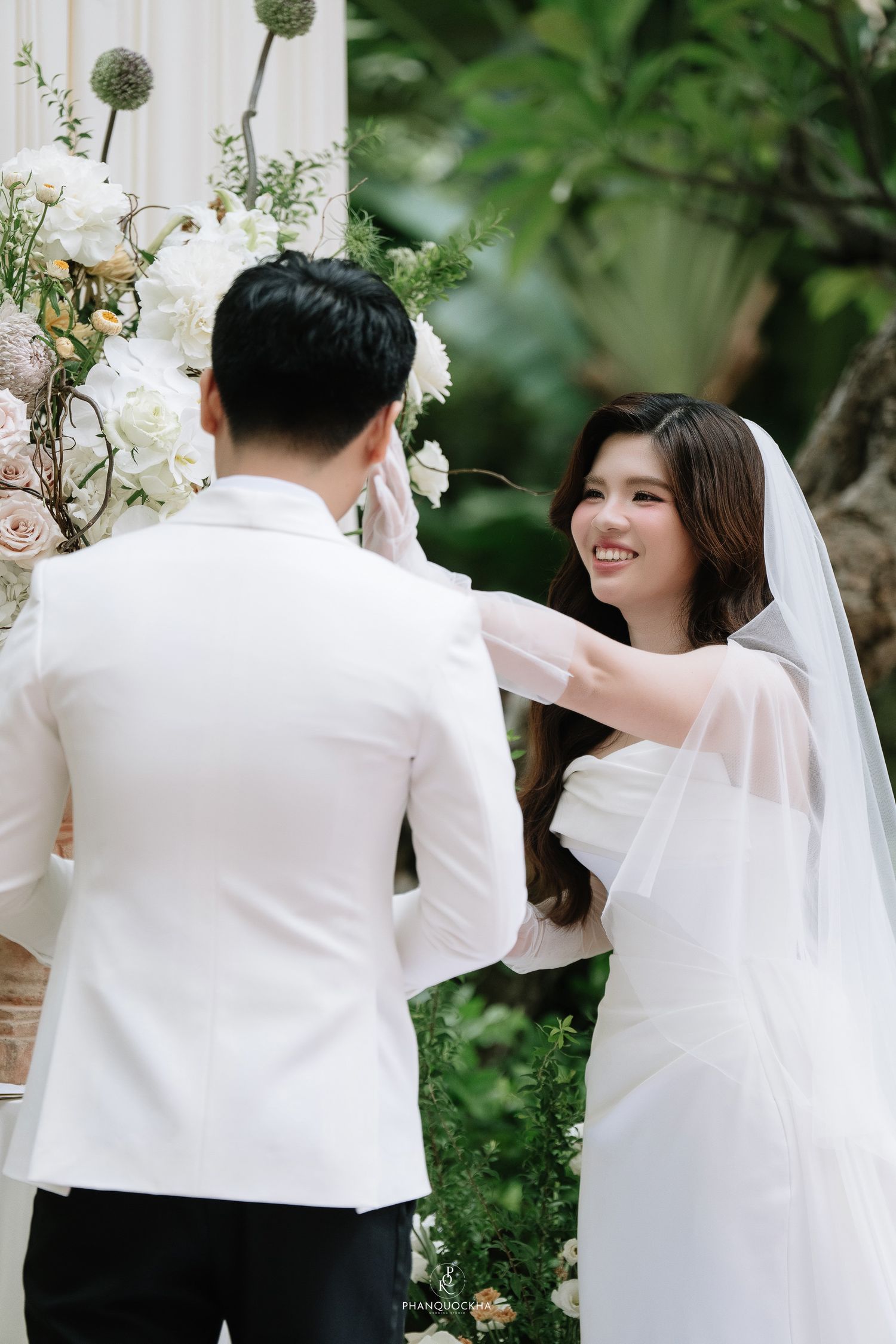 Smiling bride in white veil gently touches grooms face during outdoor wedding ceremony.