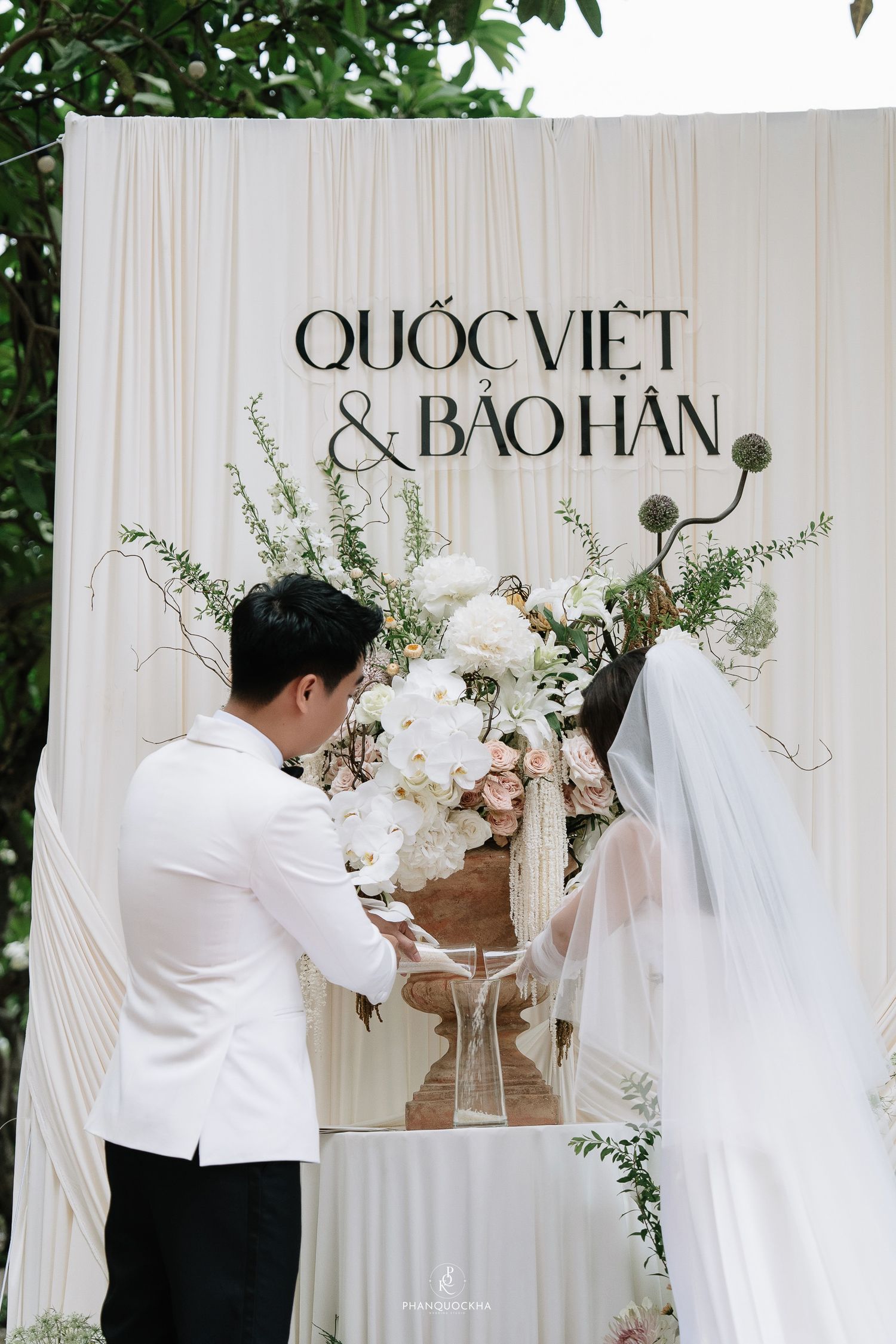 Groom and veiled bride stand before elegant Quoc Viet and Bao Han wedding backdrop with flowers.