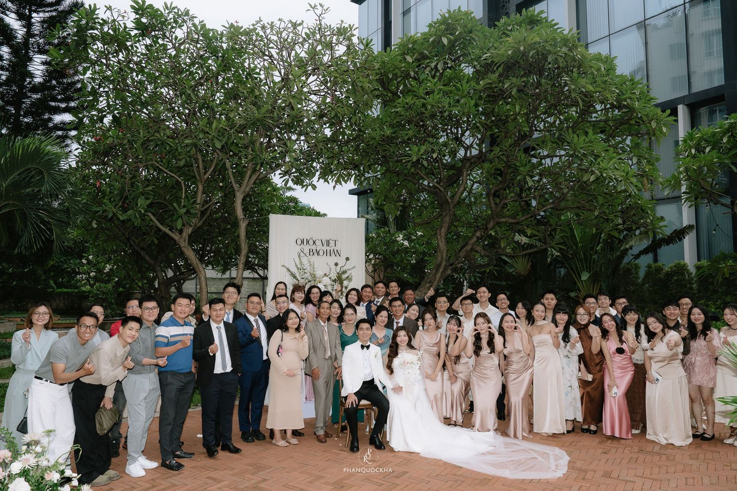 Large wedding party group photo outdoors in front of elegant venue sign surrounded by trees.