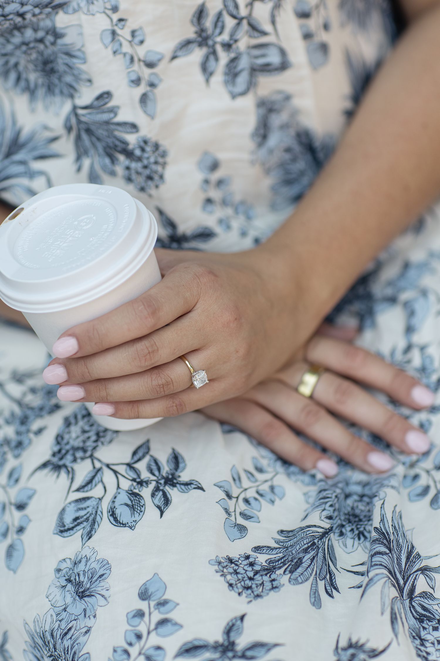 A white coffee cup held by hands wearing rings against a blue and white floral patterned fabric background.