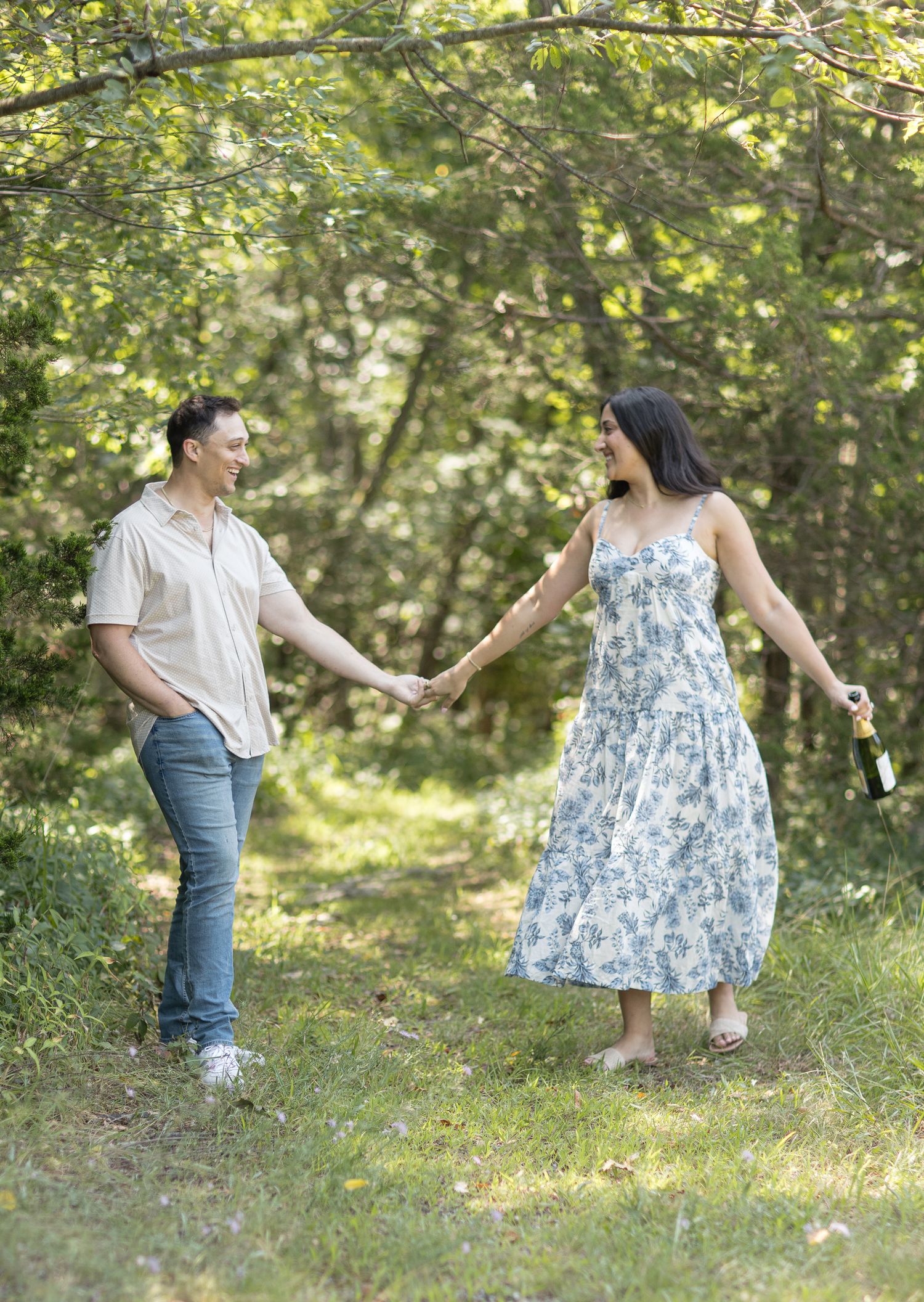 Two people hold hands while walking along a wooded path, with her wearing a floral dress and him in casual attire.