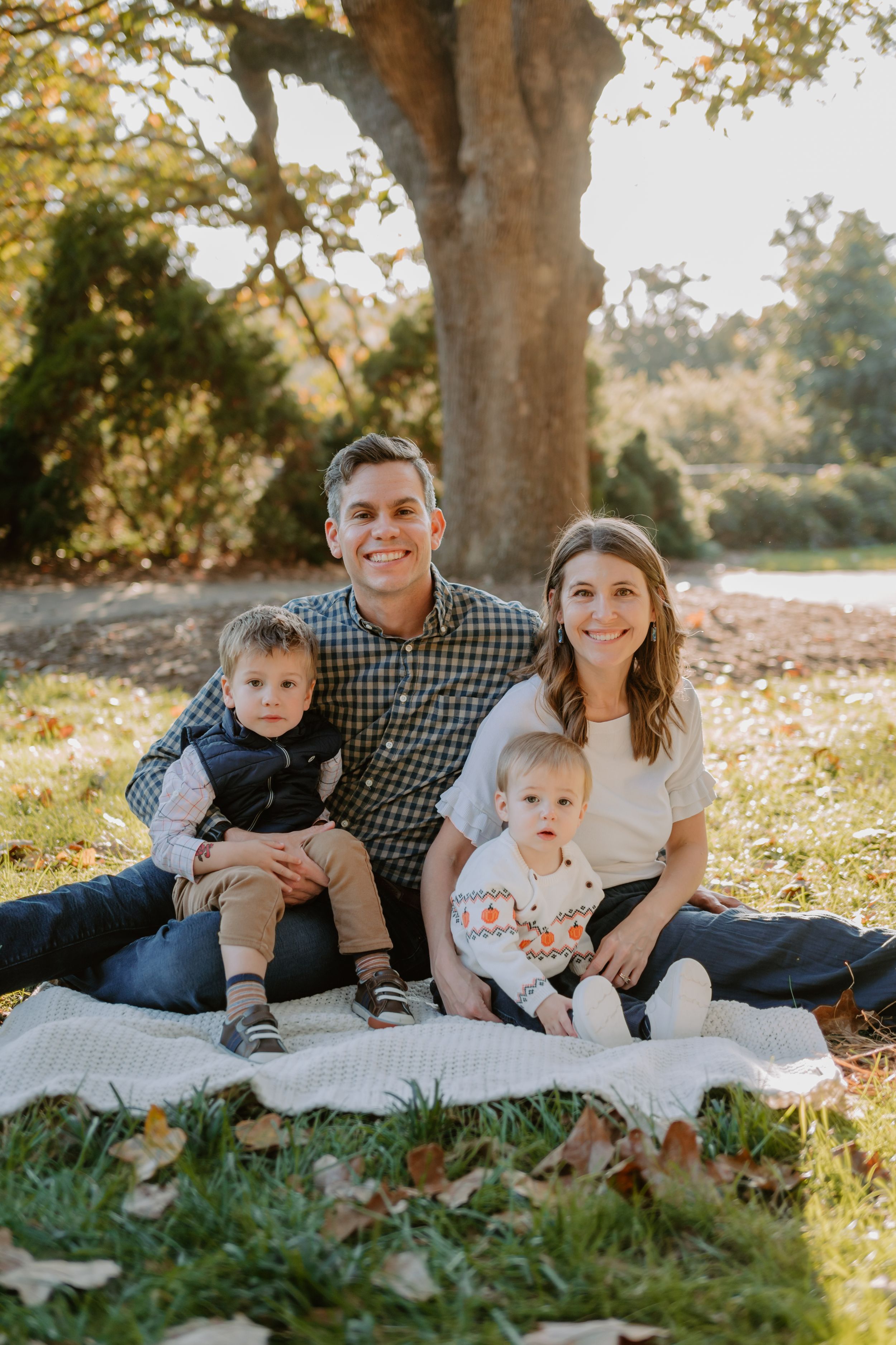A young family sits together on a white blanket under a large tree during a sunny autumn day.