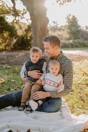 A heartwarming autumn scene shows a parent with two young children sitting on a blanket in golden sunlight.