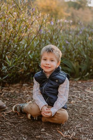 Child in navy puffer vest and plaid shirt sits cross-legged on ground during golden hour with greenery behind.