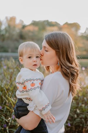 A mom in a white sweater kisses her baby wearing matching pumpkin-patterned knitwear outdoors.