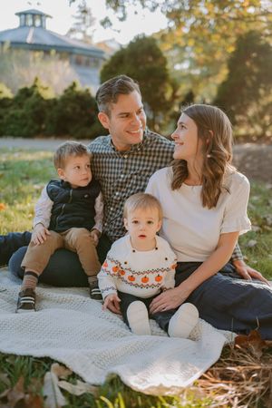 A family sits together on a white blanket during a sunny autumn day with a gazebo visible in the background.
