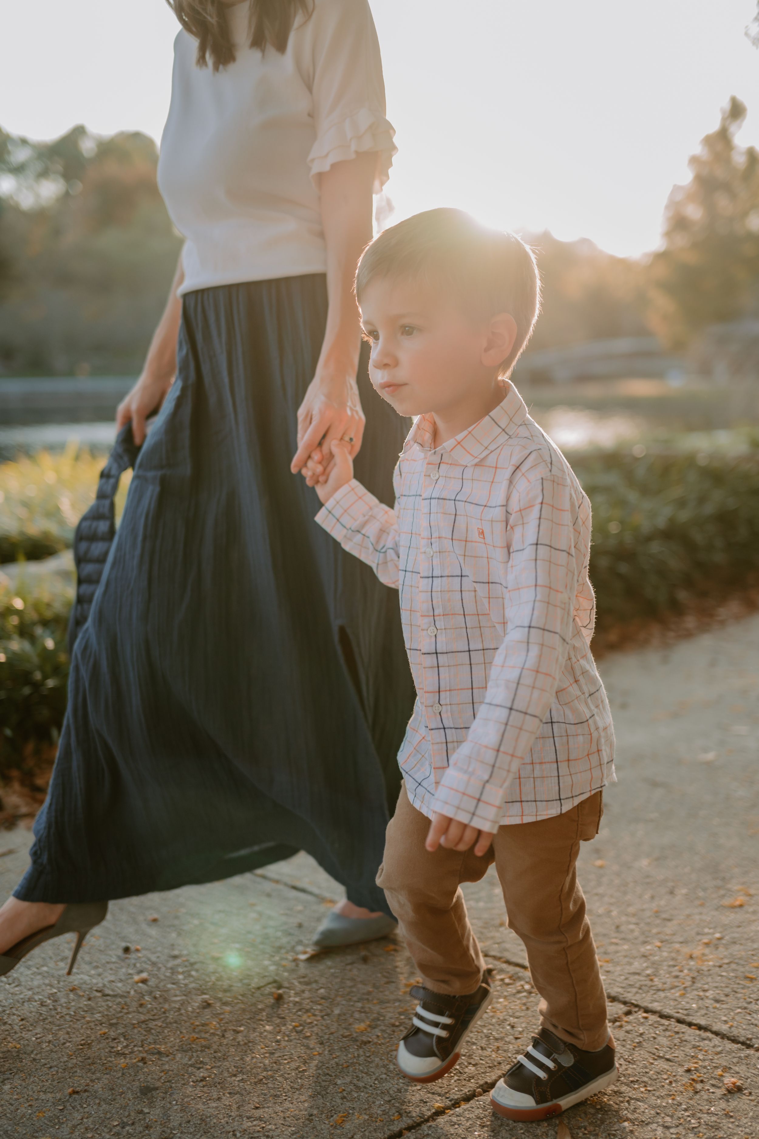 Child in plaid shirt holding hands with adult in long skirt during sunset stroll outdoors.