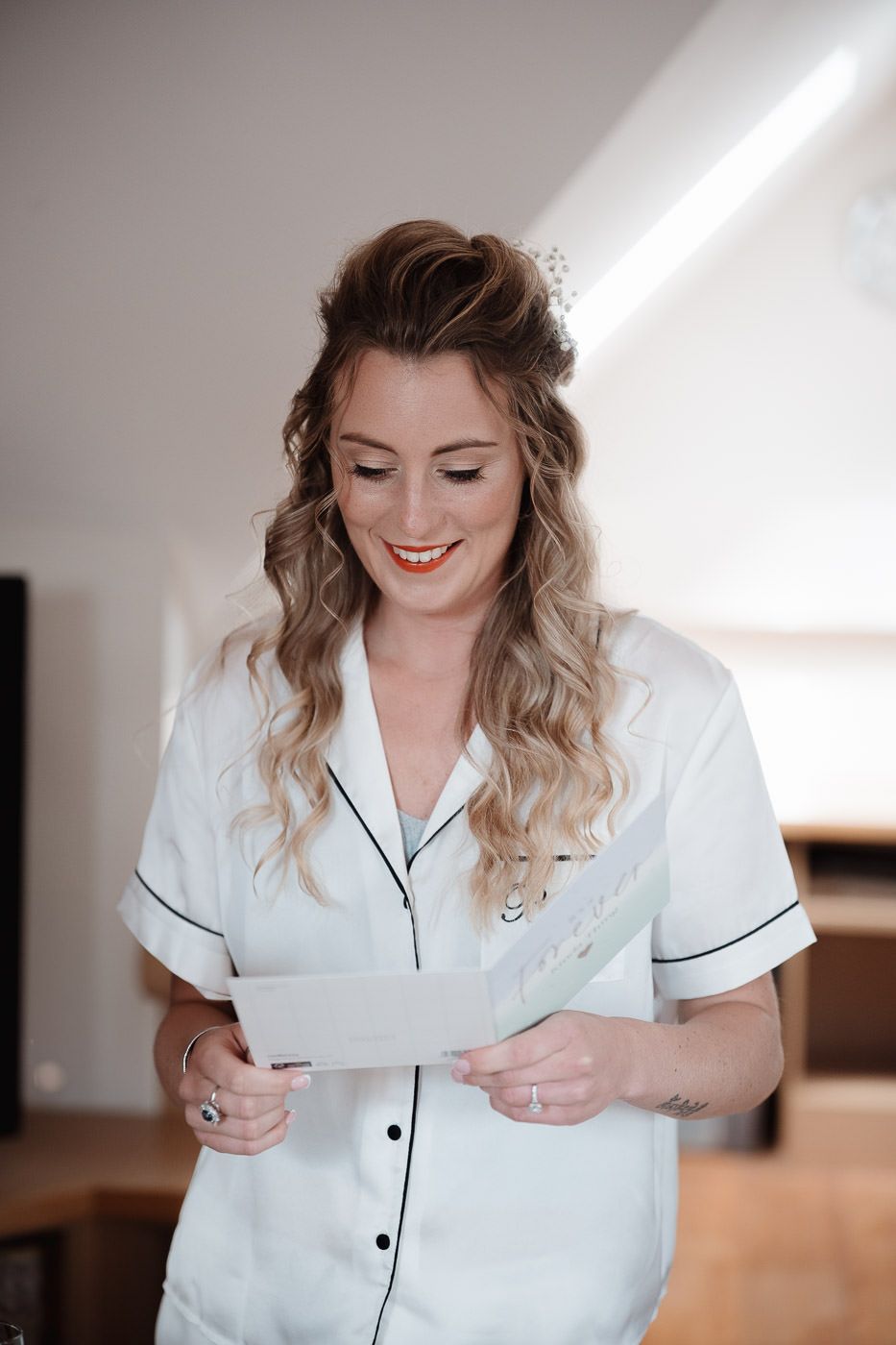 Woman in white pajama-style top smiles while reading paper in sunlit room with ombre hairstyle.