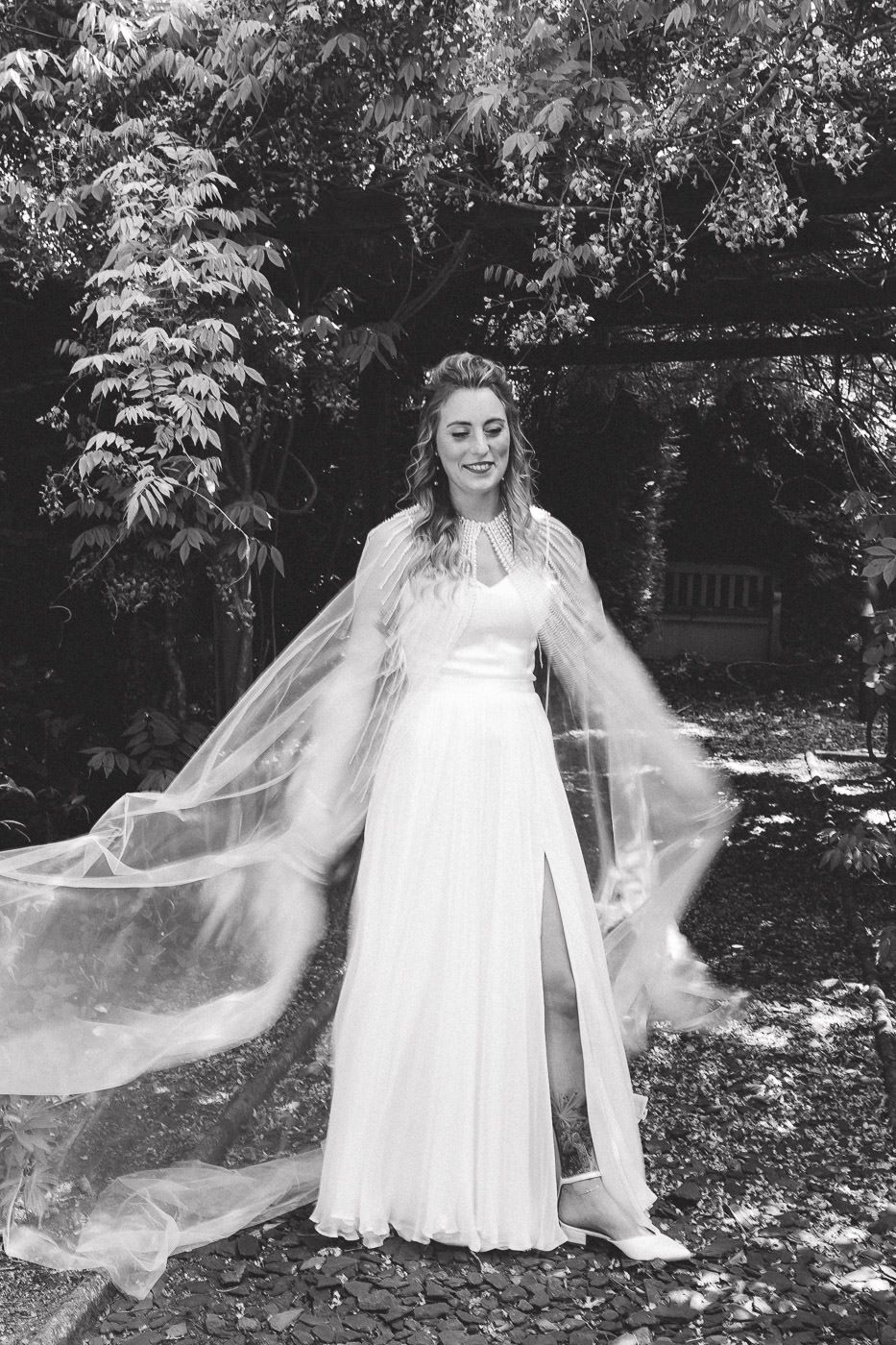 A black and white photo of a bride in a flowing wedding dress and veil standing in a garden setting.