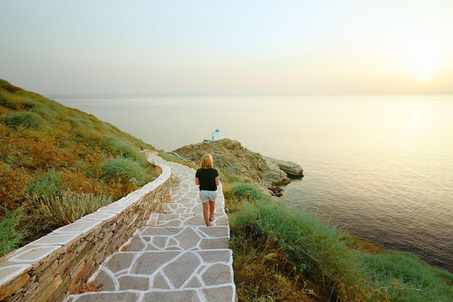 Woman walking down a stone path towards the sea at sunset