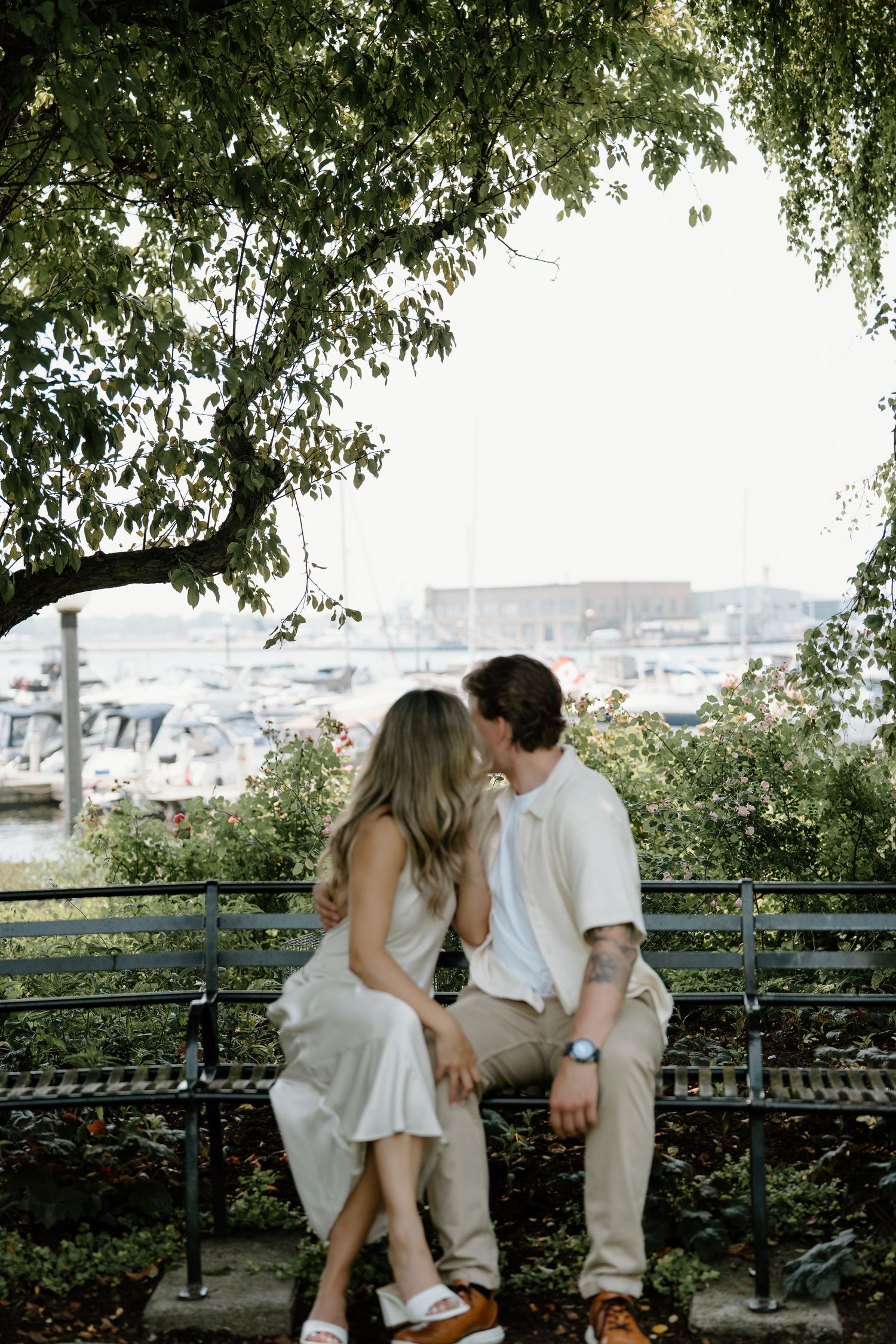 A romantic embrace on a grassy lawn near a marina with a weeping willow tree overhead.