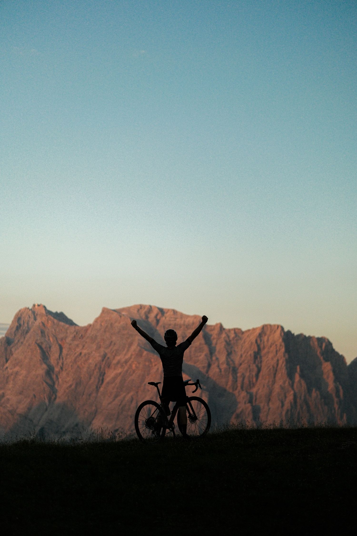 Silhouetted cyclist raises arms triumphantly against dramatic mountain backdrop at sunset.