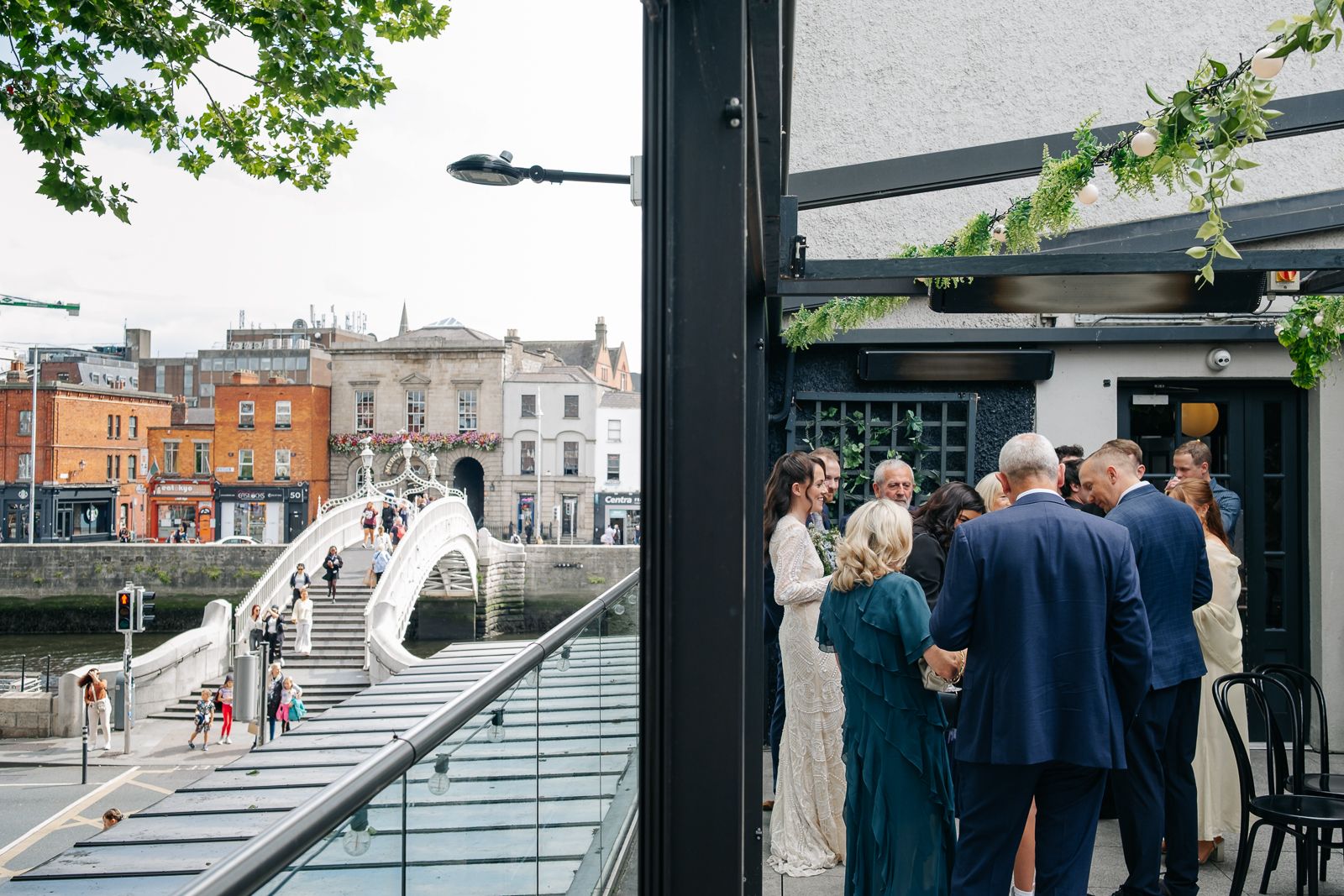 Wedding guests gather on a terrace overlooking a scenic canal bridge in an urban setting.
