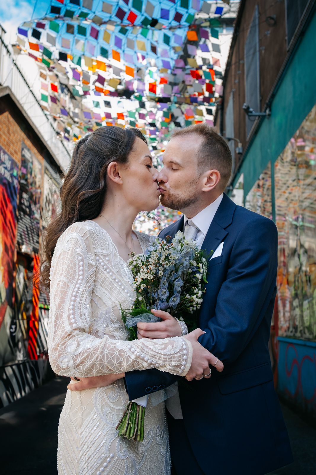 A romantic wedding couple shares a kiss under colorful hanging flags in an urban alley setting.