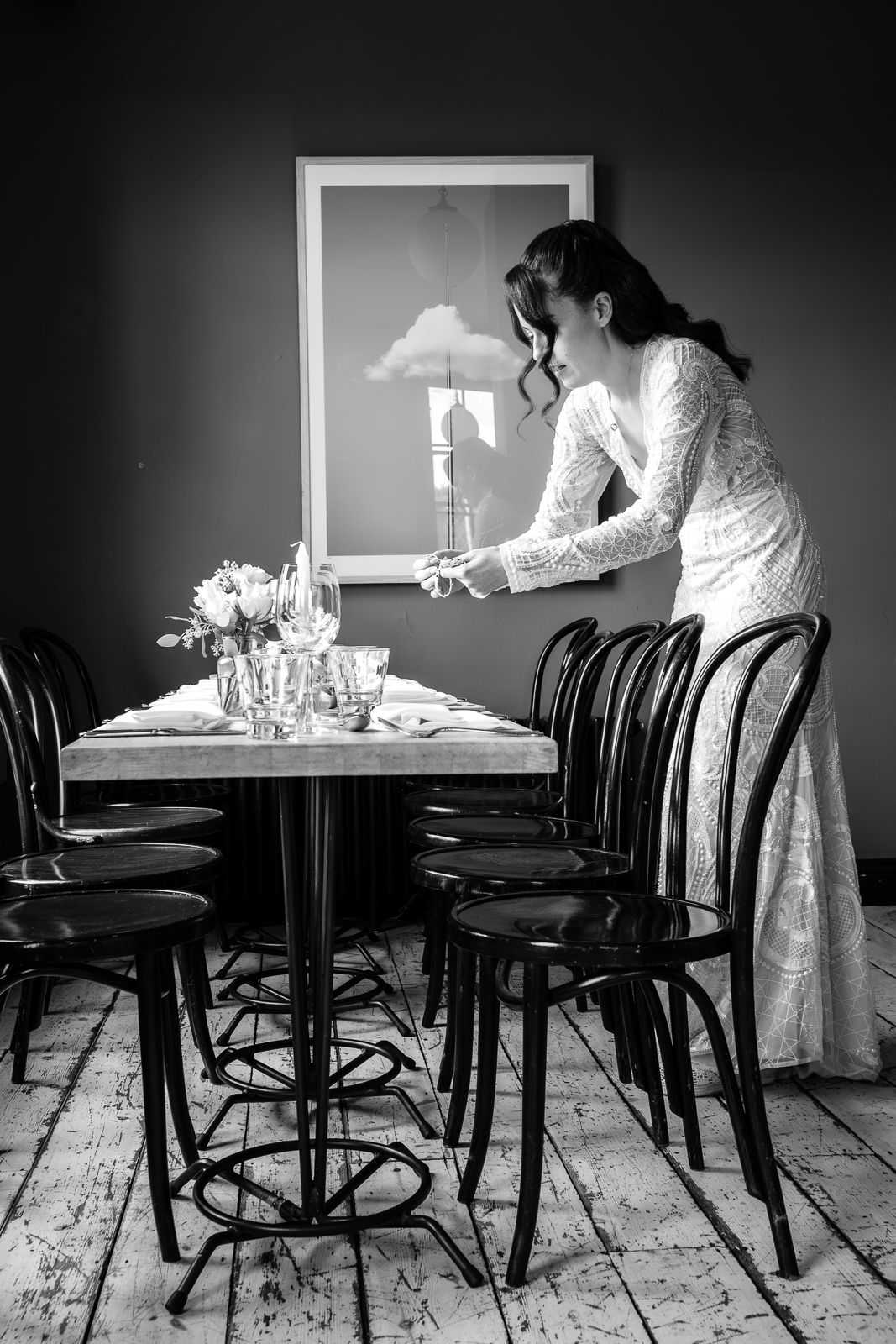 A person in a white lace dress arranges table settings in a rustic dining room with bentwood chairs.