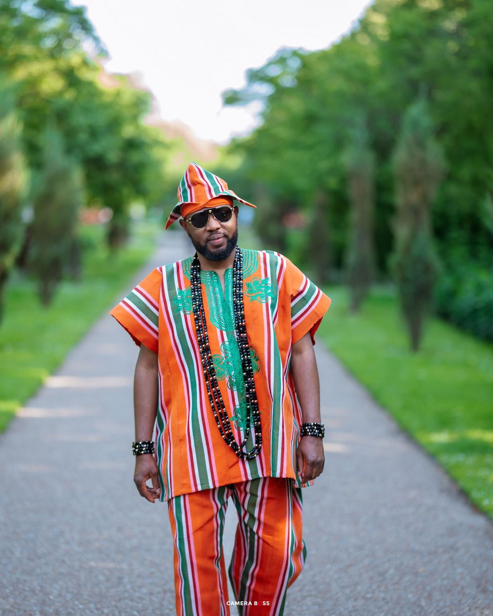 Person wearing vibrant orange striped traditional outfit with matching hat stands on paved path surrounded by greenery.
