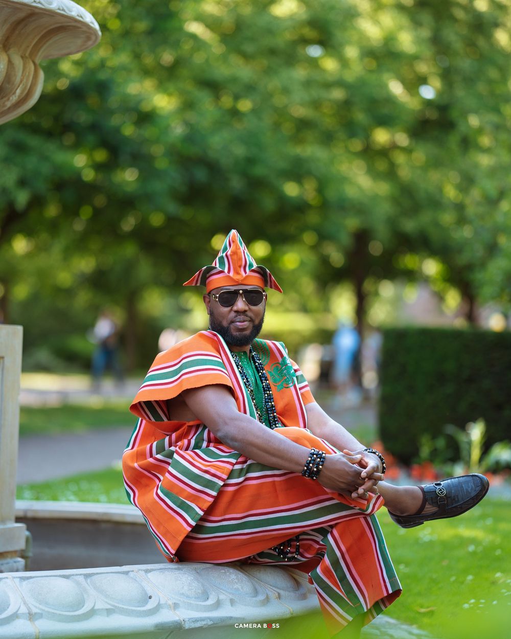 Person wearing vibrant orange traditional African attire and sunglasses sits casually in an outdoor park setting.
