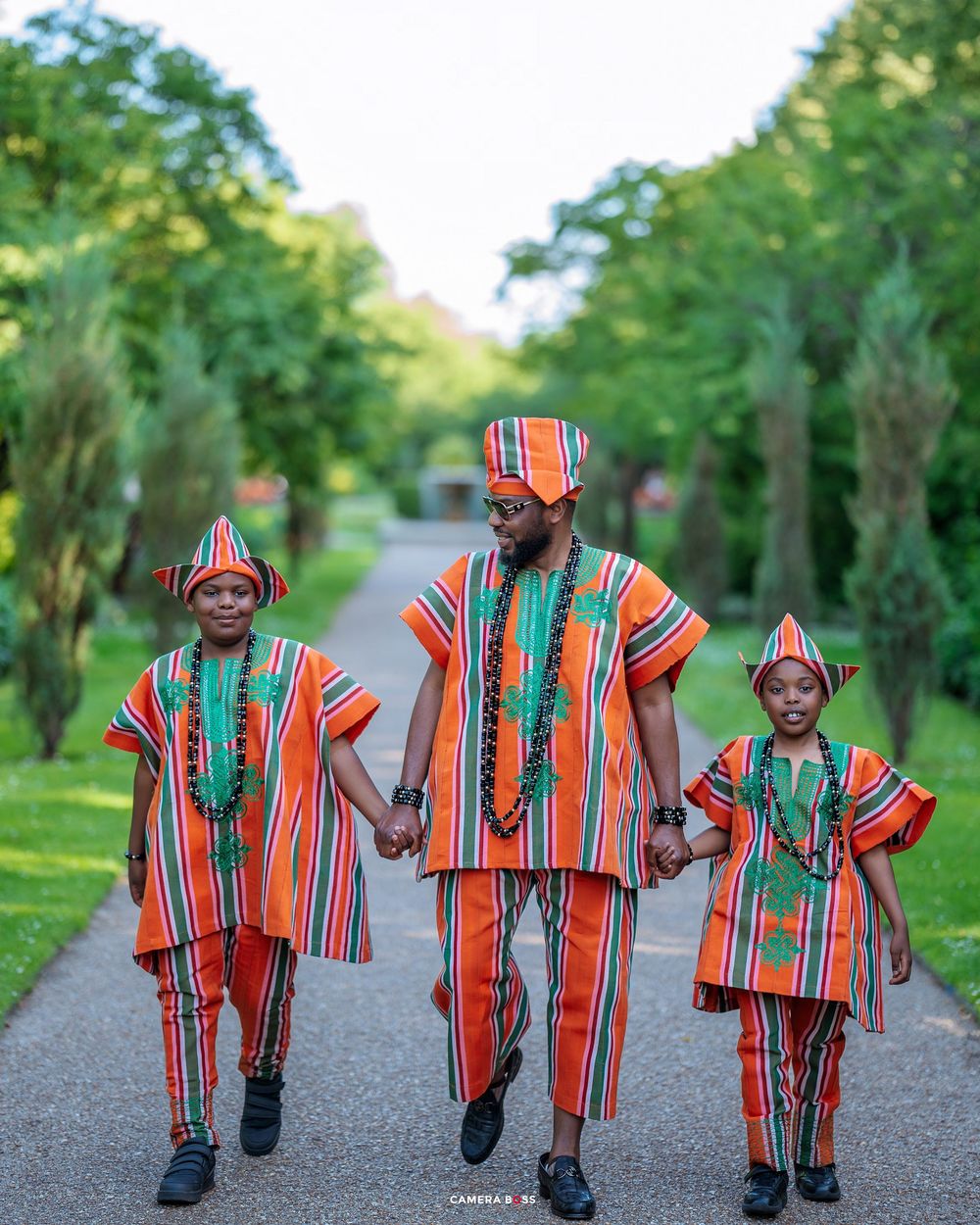 Three people walk hand in hand wearing vibrant orange and green striped traditional African outfits on a tree-lined path.