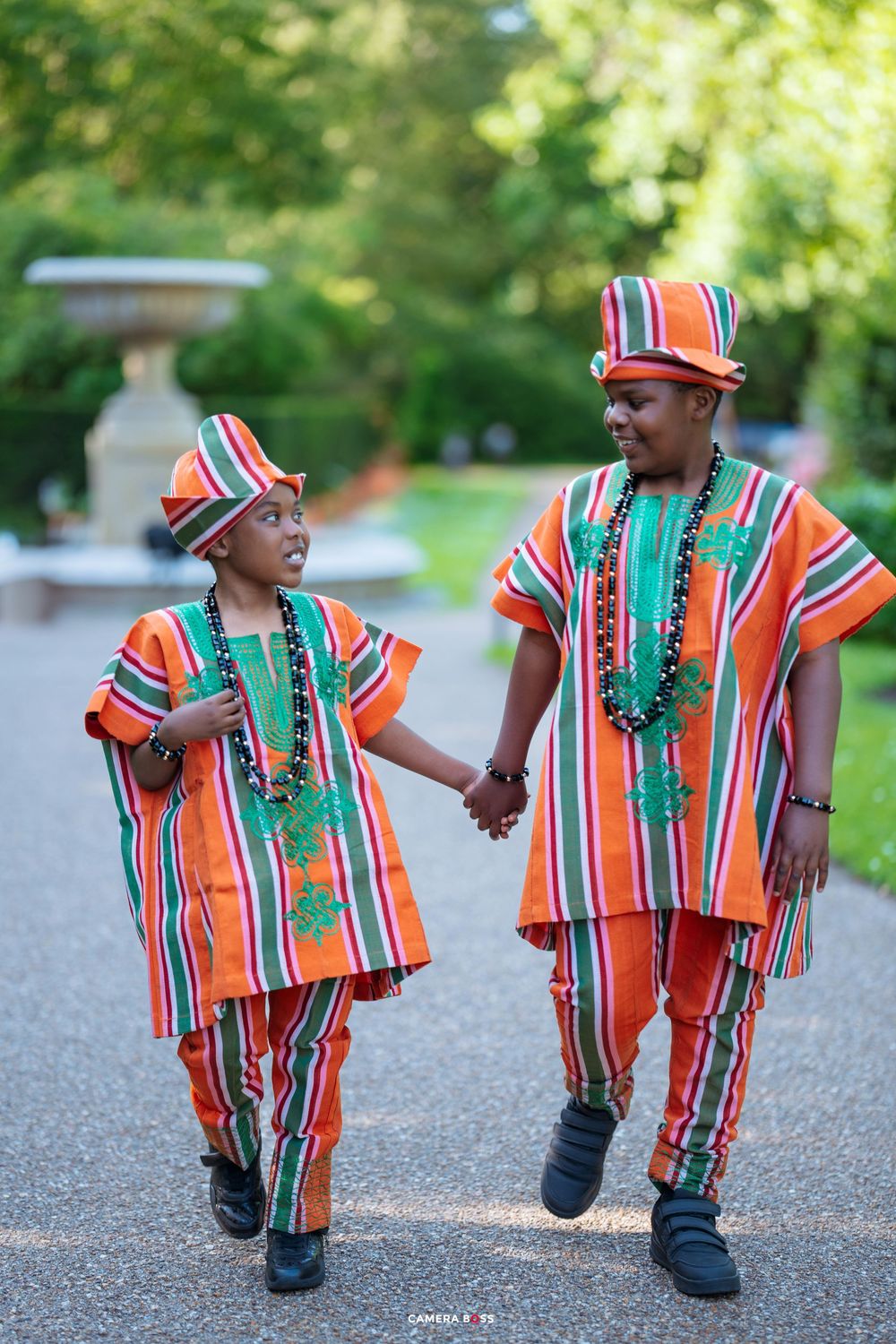 Two people wearing matching orange and white striped traditional outfits with green beaded necklaces walk hand in hand outdoors.