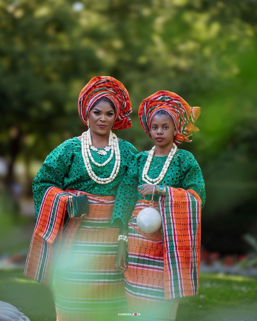 Two people wearing traditional African attire with colorful headwraps, green lace tops and striped wrappers pose in a garden setting.