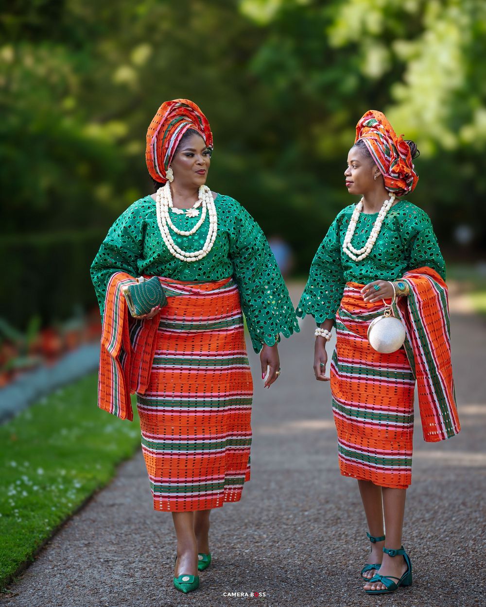 Two women in vibrant green and orange African traditional attire with gele headwraps walk along a garden path.