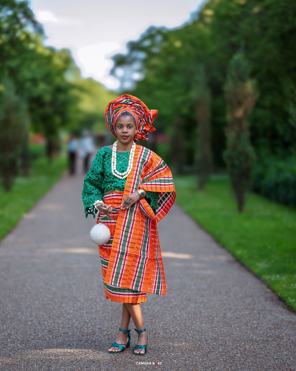 Traditional African fashion featuring orange plaid wrapper, green top and matching headwrap on garden pathway.