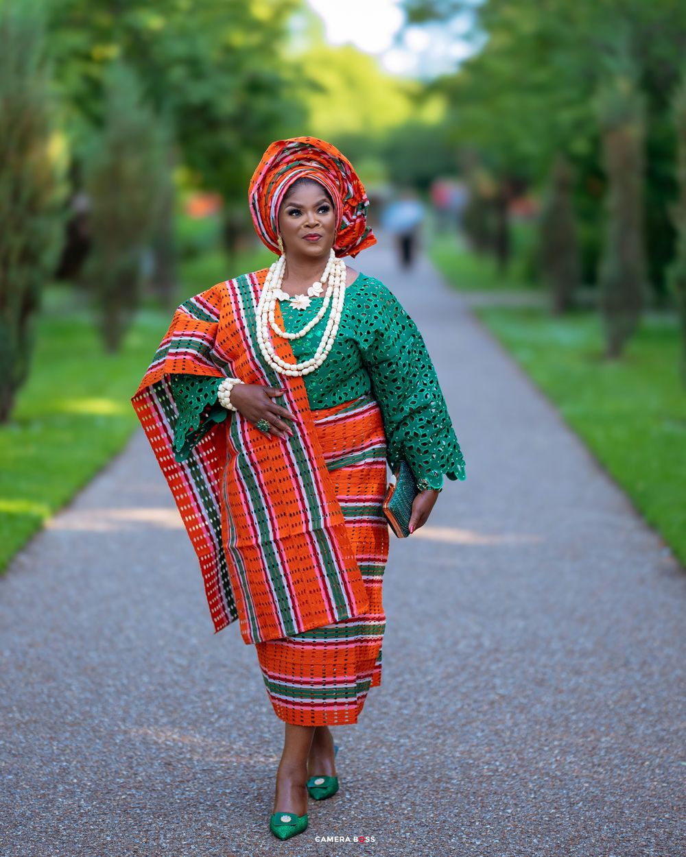 African traditional outfit with emerald lace top, orange plaid wrapper and matching gele on park pathway.