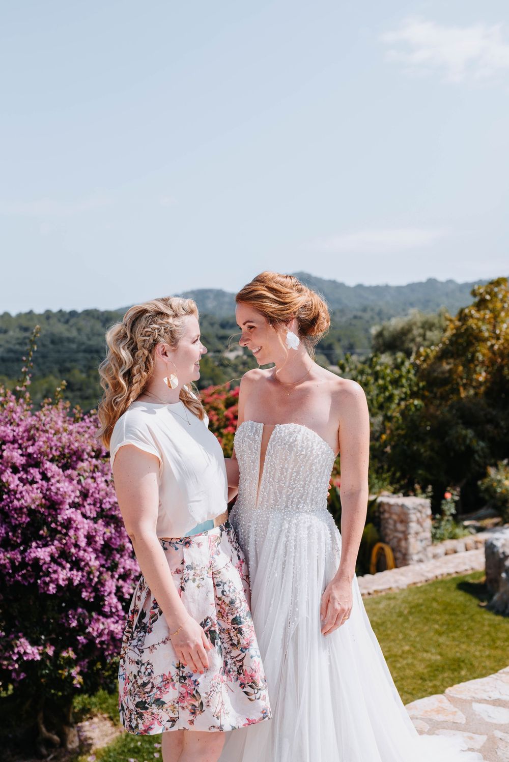Braut und ihre Schwester schauen sich, auf einer malerischen Terrasse mit blühenden rosa Blumen und Bergblick Mallorcas im Hintergrund, an.