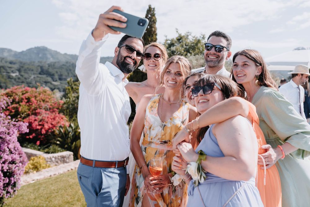 Hochzeitsgäste machen mit dem Brautpaar ein lachendes Selfie an der malerischen Finca Xiclati im Freien mit Blick auf die Berge Mallorcas.
