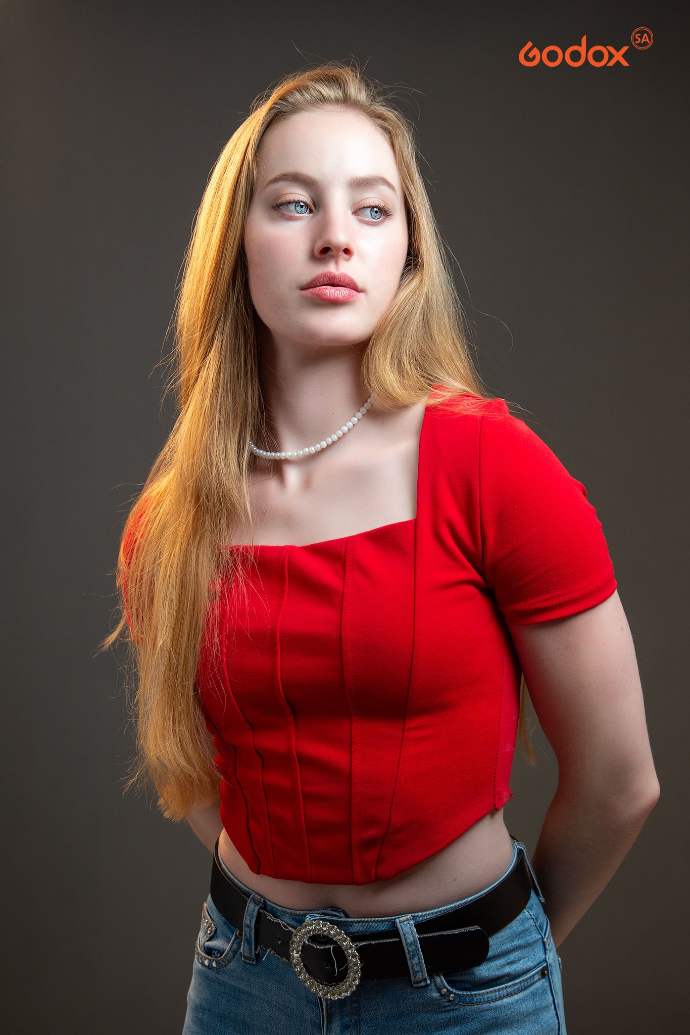 Studio portrait featuring a red cropped top with short sleeves paired with blue jeans and a decorative belt on dark background.