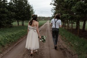 A couple walks down a tree-lined dirt path, with the bride carrying a white bouquet.