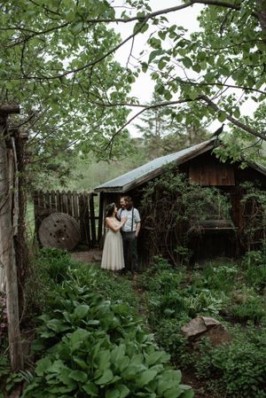 A couple stands in a lush garden setting beside a rustic wooden cabin with green foliage.