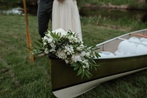 White floral bouquet resting on a wooden canoe with wedding attire visible in background.