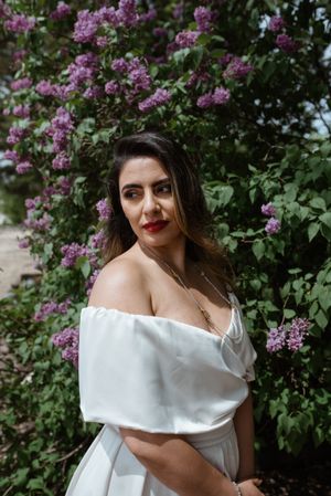 Off-shoulder white dress against blooming lilac bush in garden setting.