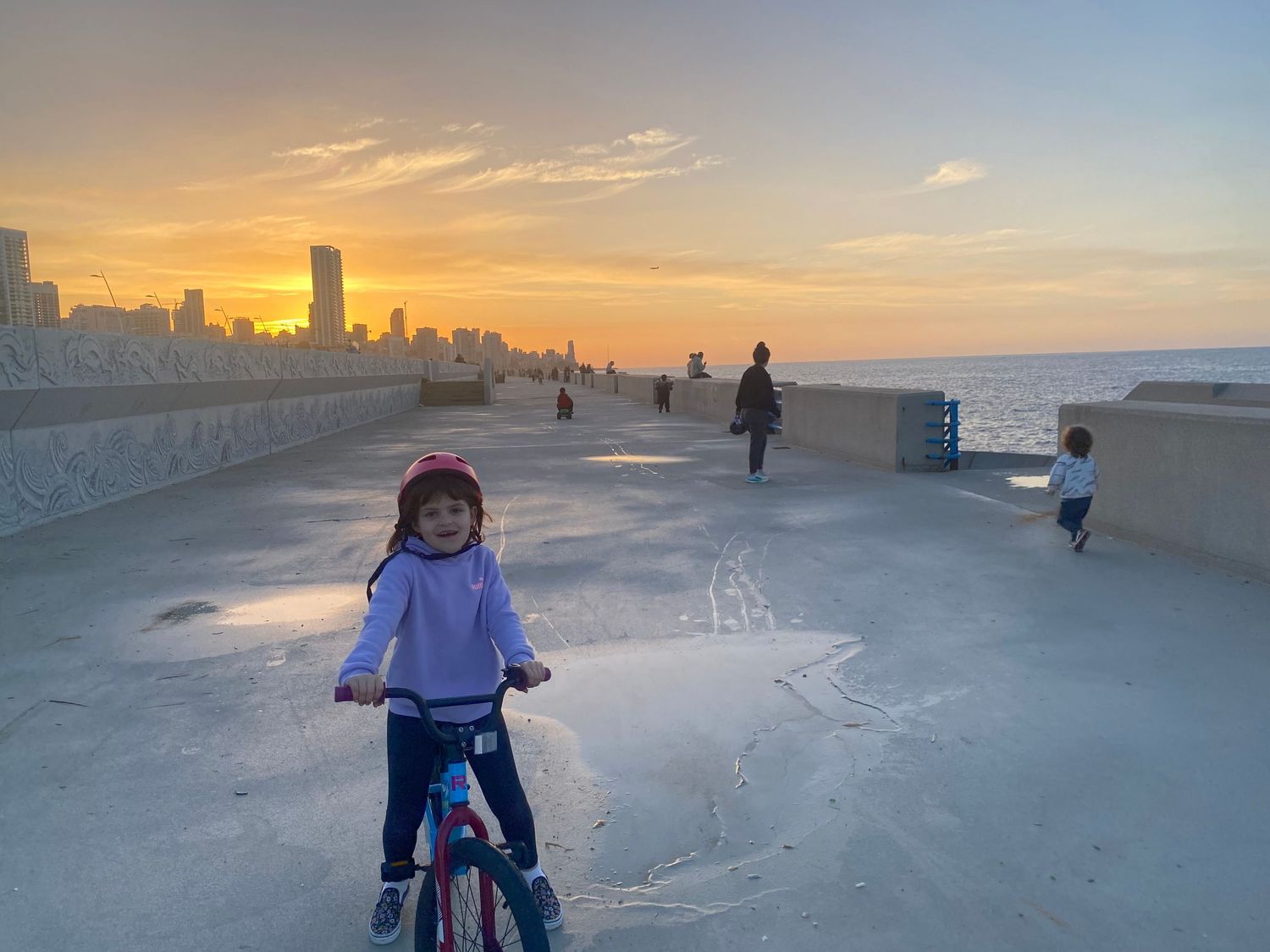 Person riding a bike along a beachfront promenade at sunset with city skyline in the background.