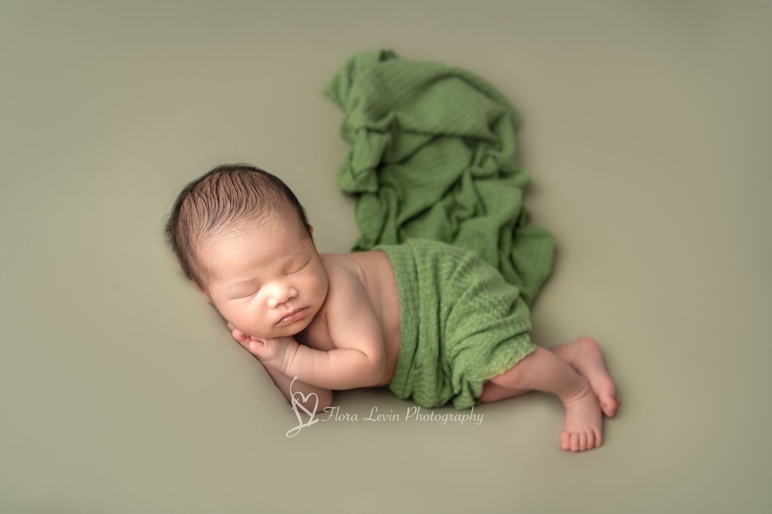 Sleeping newborn baby wrapped in soft green fabric lying on matching green background for professional portrait.