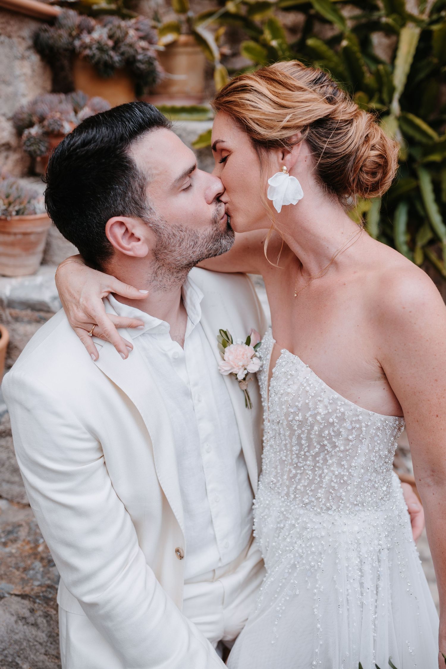Romantisches Hochzeitspaar in passenden weißen Outfits, das sich vor einer rustikalen Steinmauer mit grünen Pflanzen innig küsst.