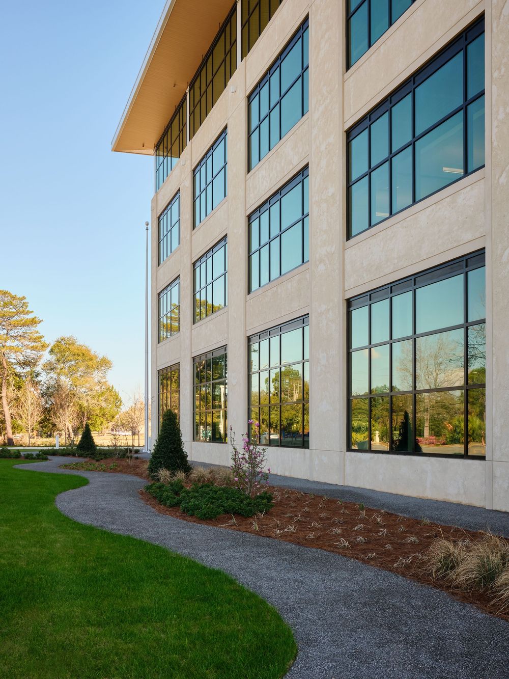 Modern office building exterior with large windows and curved walkway surrounded by landscaped lawn and trees.