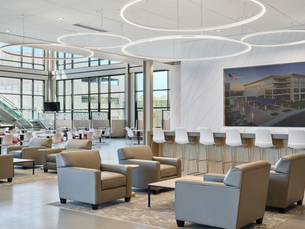 Modern lobby waiting area with circular ceiling lights, comfortable tan and gray armchairs, and large windows overlooking outdoors.