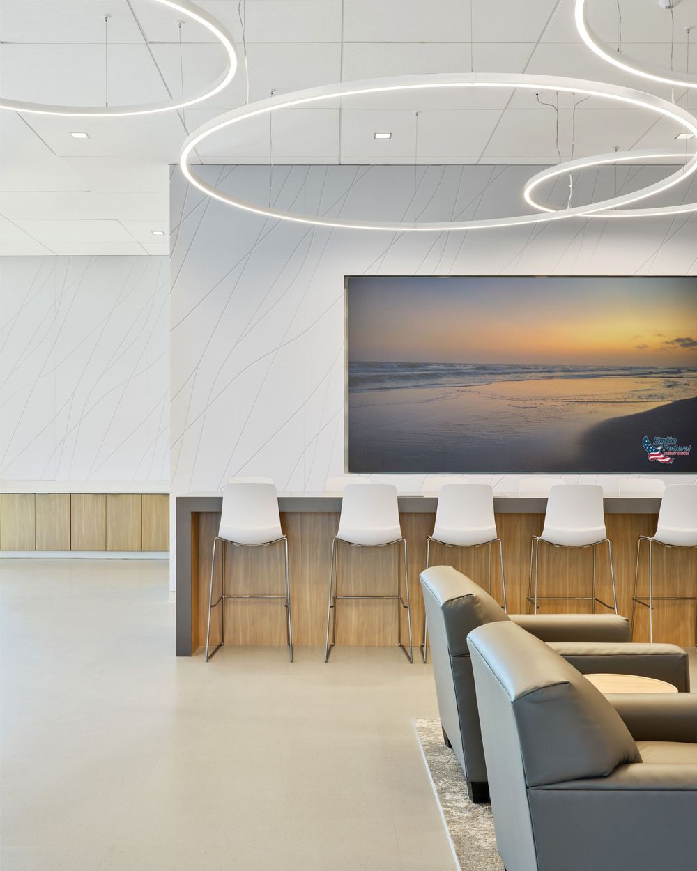 Modern office lounge with curved LED lighting fixtures, bar seating, and a scenic beach sunset photograph on the wall.