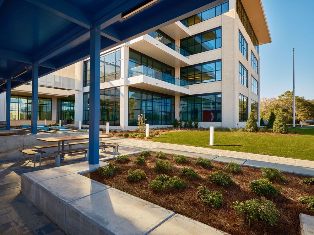 Contemporary office building exterior with tiered balconies, blue support columns, and landscaped garden areas.