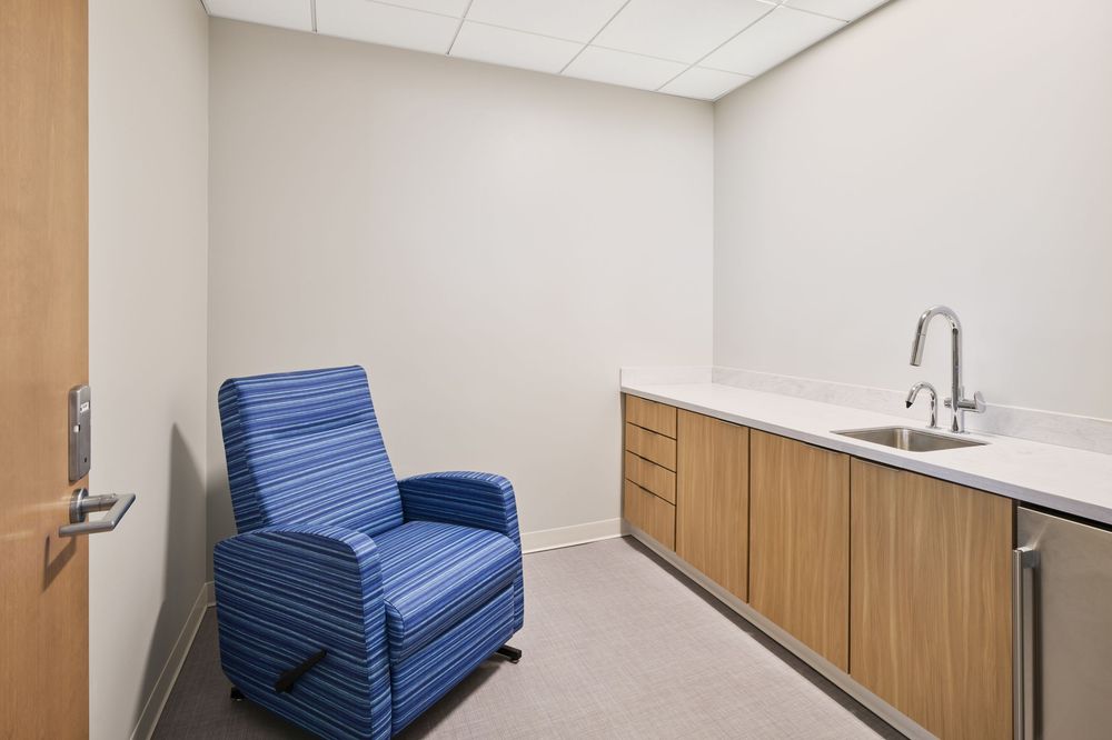 Small medical room with blue striped chair, wood cabinets, and white countertop with sink.