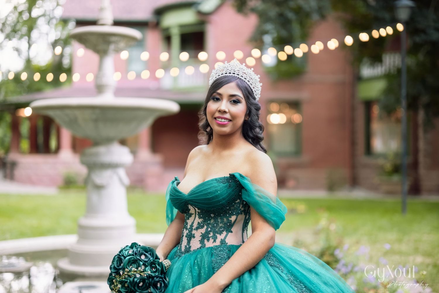 A person in a stunning emerald green off-shoulder ball gown poses in front of a pink building with string lights.