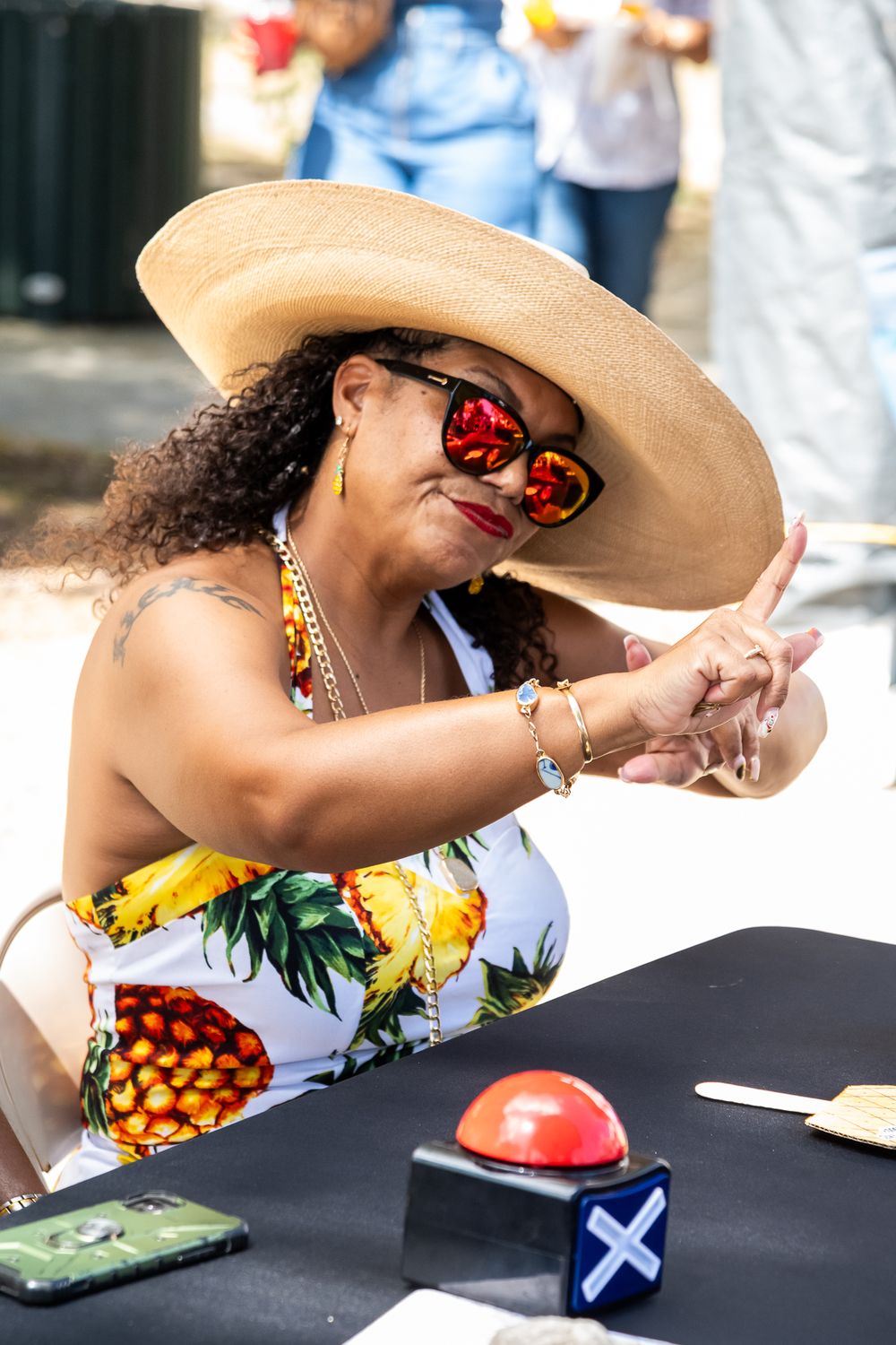 A festival-goer in a wide-brimmed hat, tropical print top and mirrored sunglasses smiles while seated at a table.