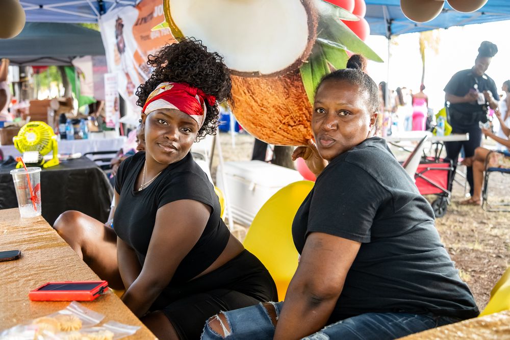 Two people in black shirts sitting together at a festival with tropical decorations visible in the background.