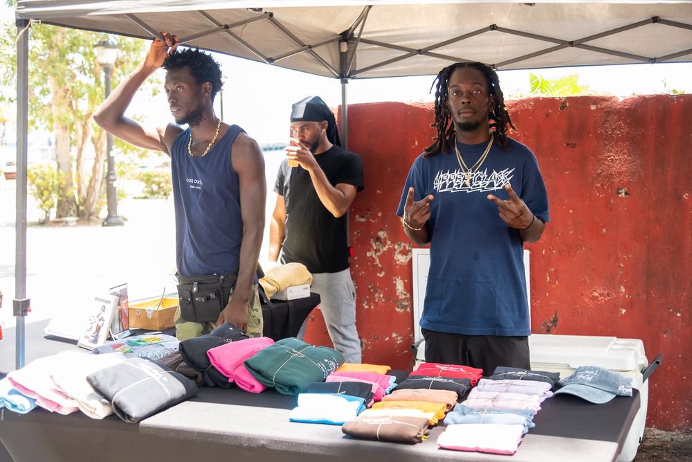 Three people staffing a merchandise table with folded t-shirts and apparel displayed under a white canopy tent.