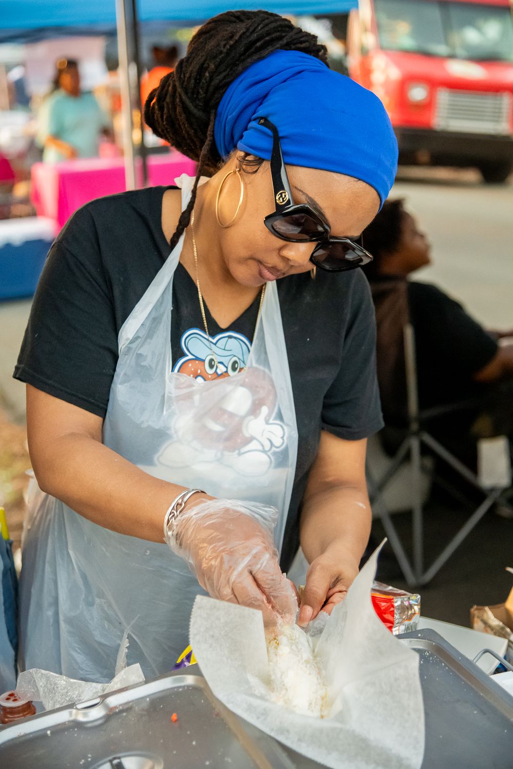 A vendor in a blue headband and apron prepares food at an outdoor market stall.
