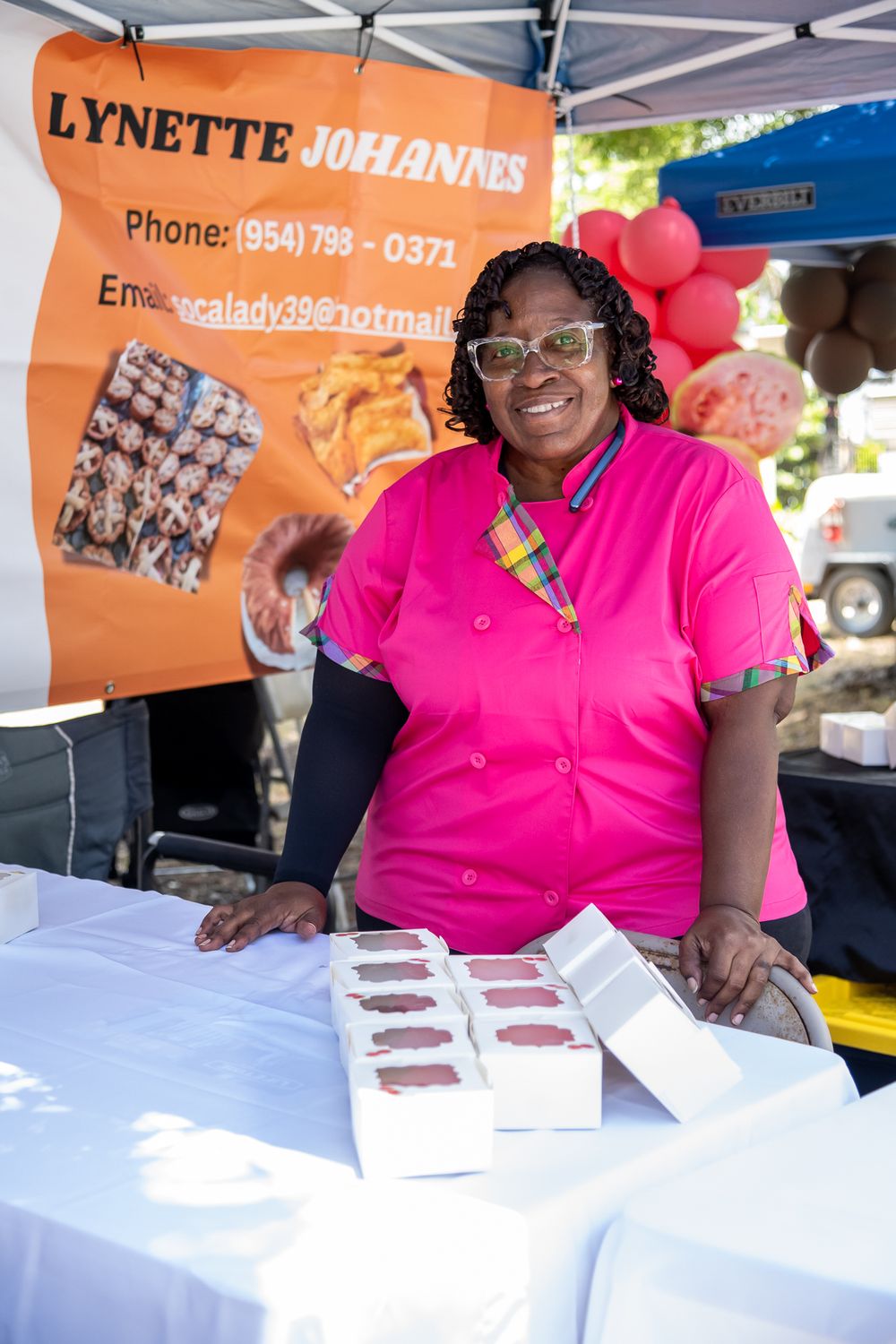 A vendor wearing bright pink sits at a table with business cards and promotional materials at an outdoor event.