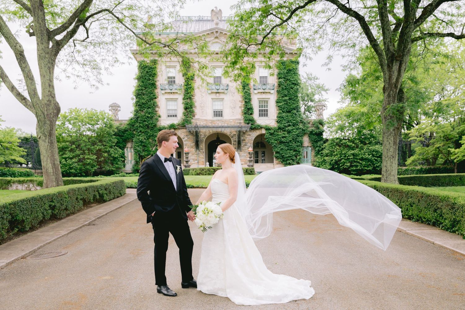A wedding couple poses in front of an ivy-covered mansion with the bride's veil flowing dramatically in the wind.