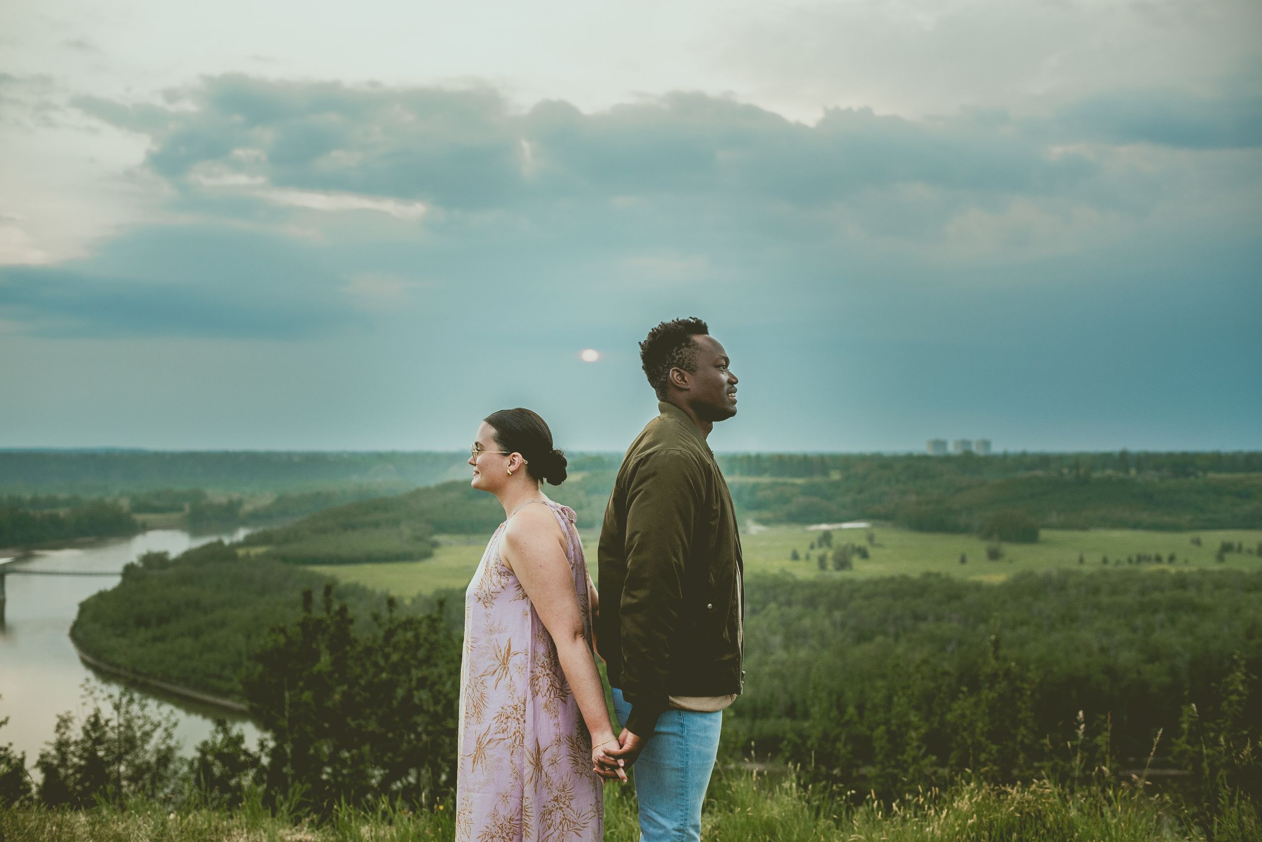 Two people stand back-to-back on a hilltop overlooking a winding river and green valley landscape at dusk.